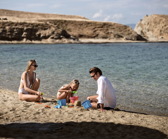 Family building sandcastles on a quiet beach by clear water, with rocky headland in the background at Drenia island
